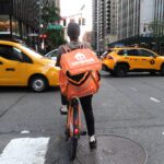 A delivery person wearing an orange Grubhub bag is seen from behind as he waits for several taxis to pass his bike.