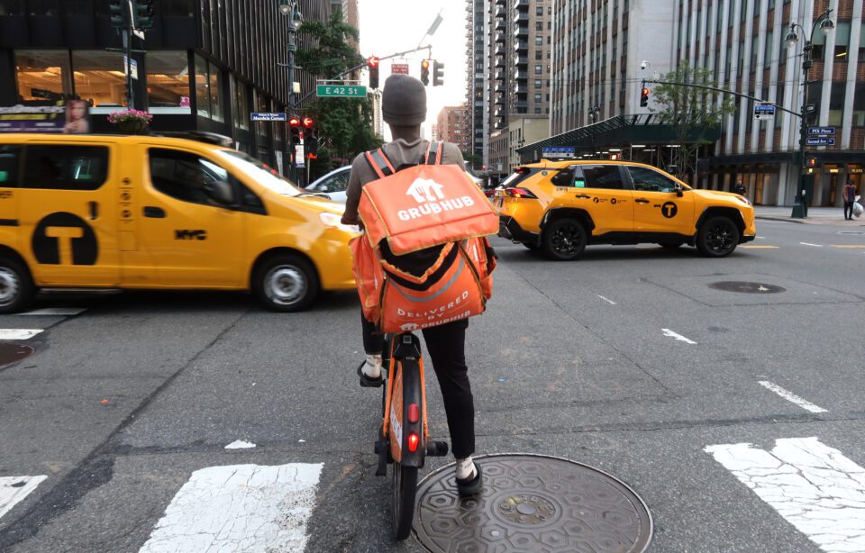 A delivery person wearing an orange Grubhub bag is seen from behind as he waits for several taxis to pass his bike.