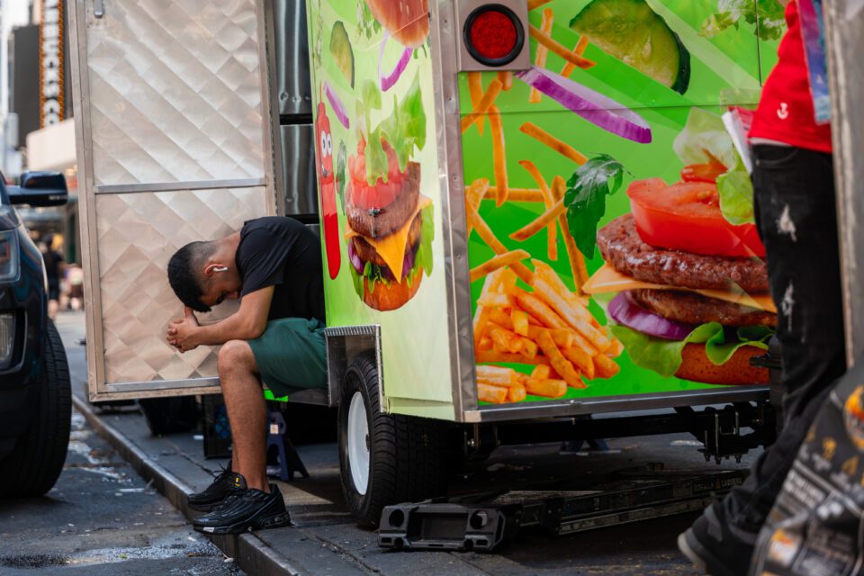 A delivery person sits with his head down and his hands clasped in sweltering heat. He is wearing green shorts and a black shirt.
