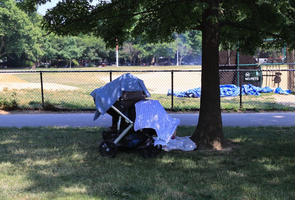 A baby stroller under the shade of a tree in a park with cloths over it to keep the baby cool in hot weather.