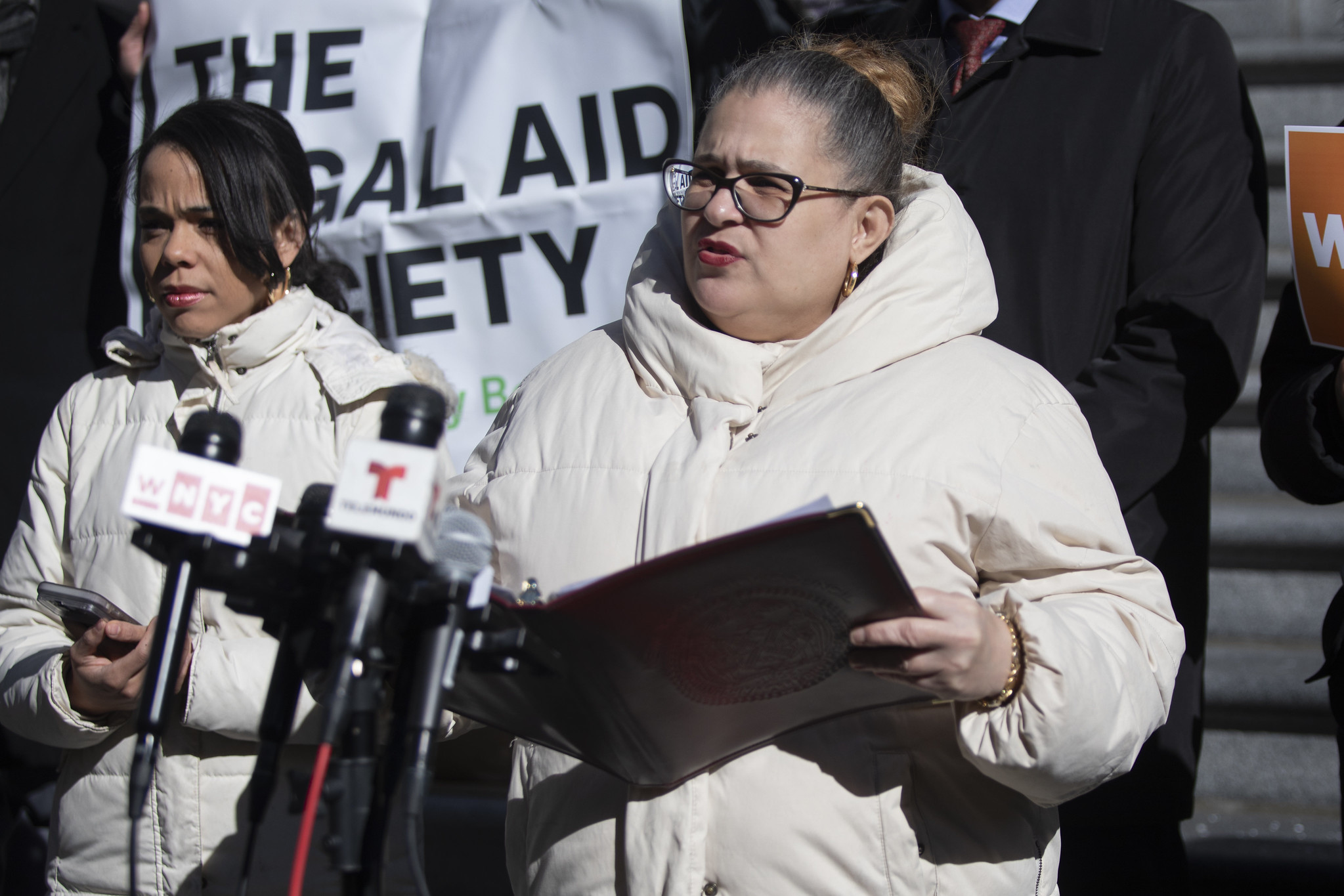 Councilmember Diana Ayala, in a cream winter puffer, speaks into microphones as a Legal Aid Society banner is shown behind her.