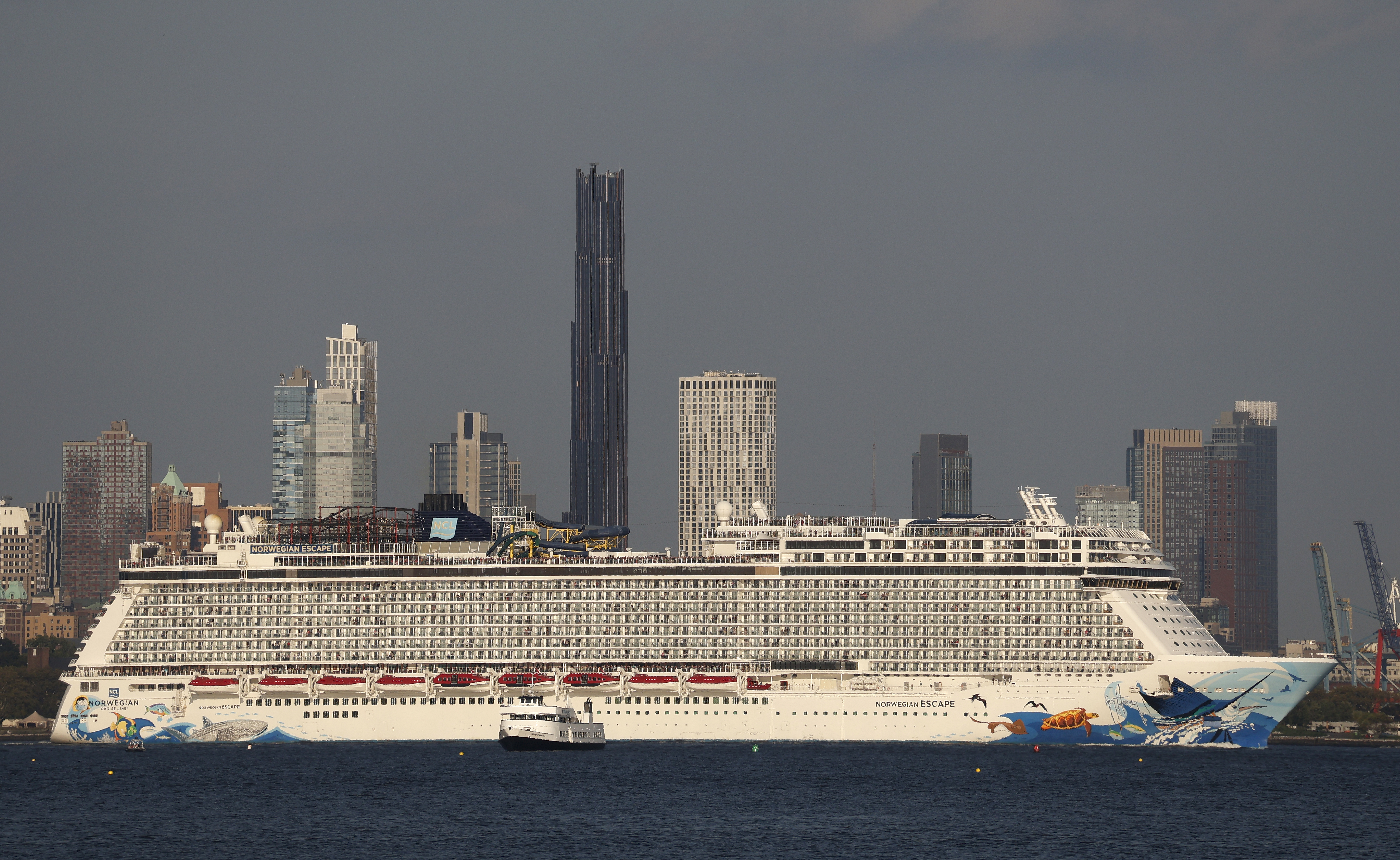 The Norwegian Escape cruise ship sails in front of the skyline of Brooklyn and the Brooklyn Tower in New York City.