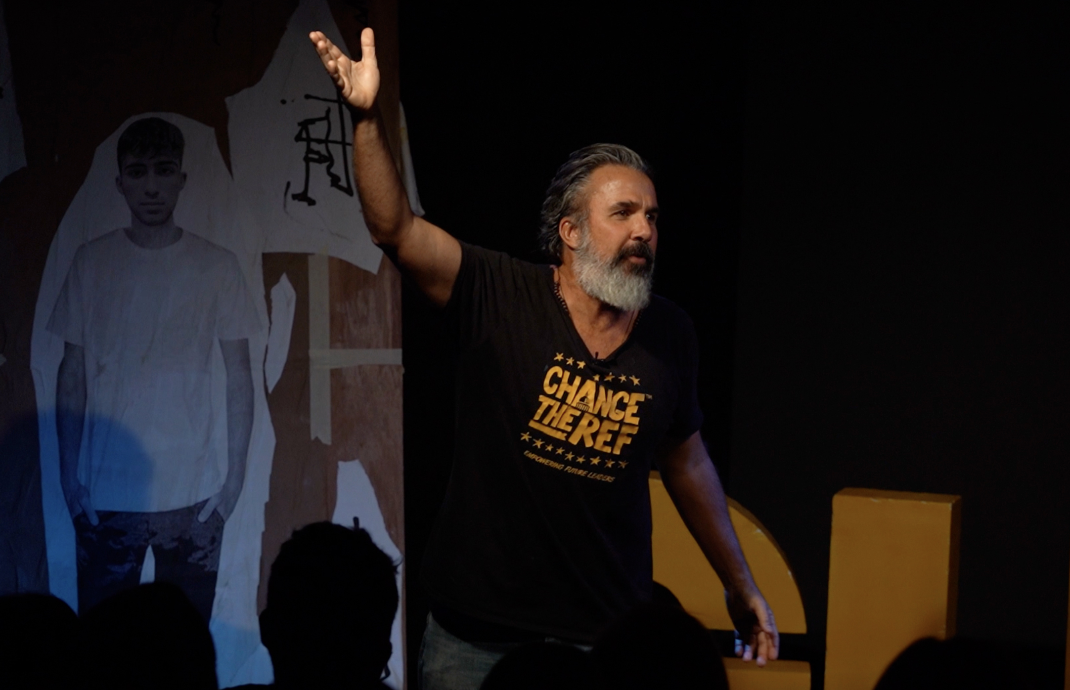 A man stands on a stage wearing a black T-shirt.