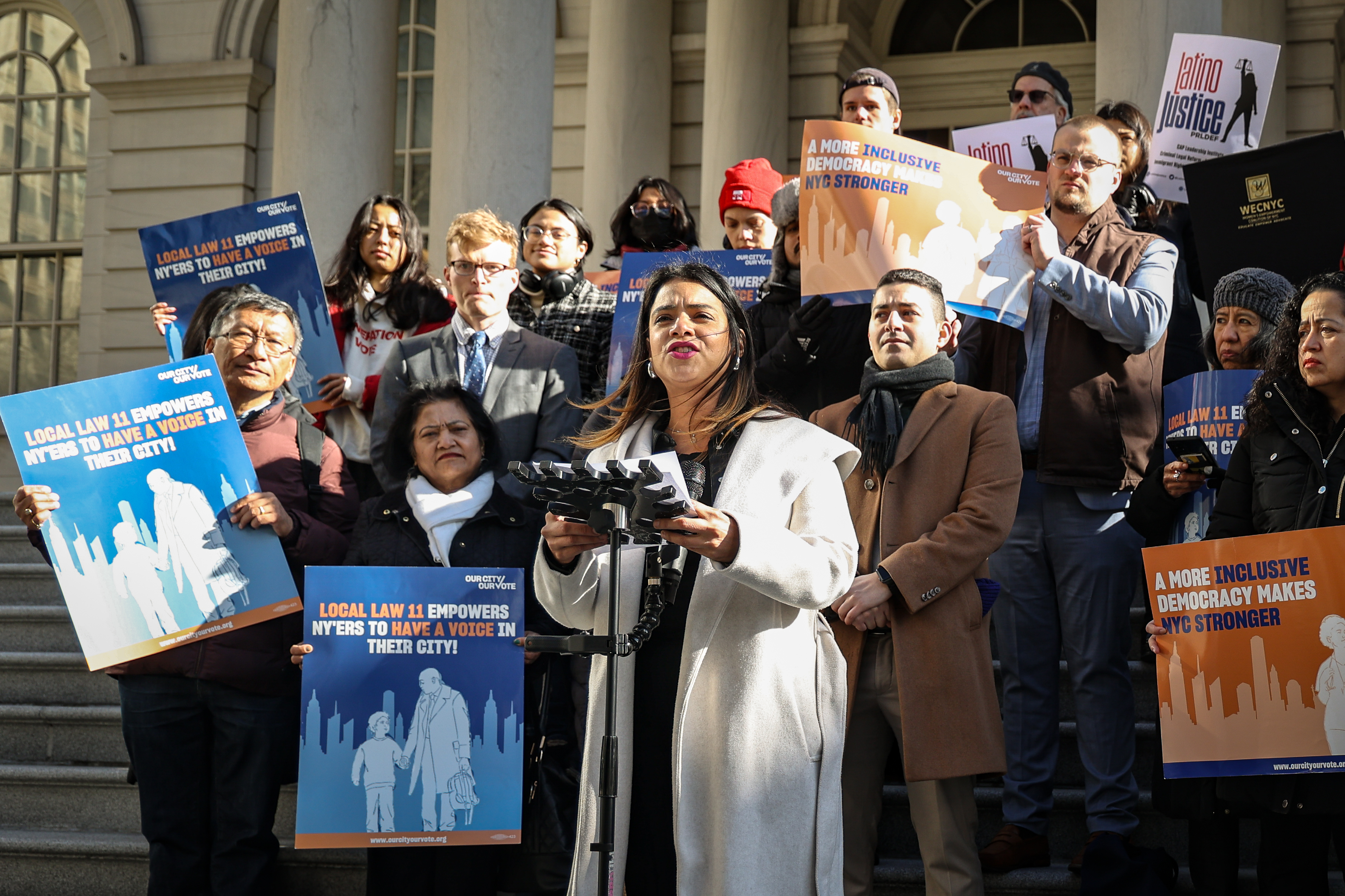 Eva Santos Veloz, in a white coat, speaks on the steps of City Hall flanked by demonstrators holding signs in favor of the local law.