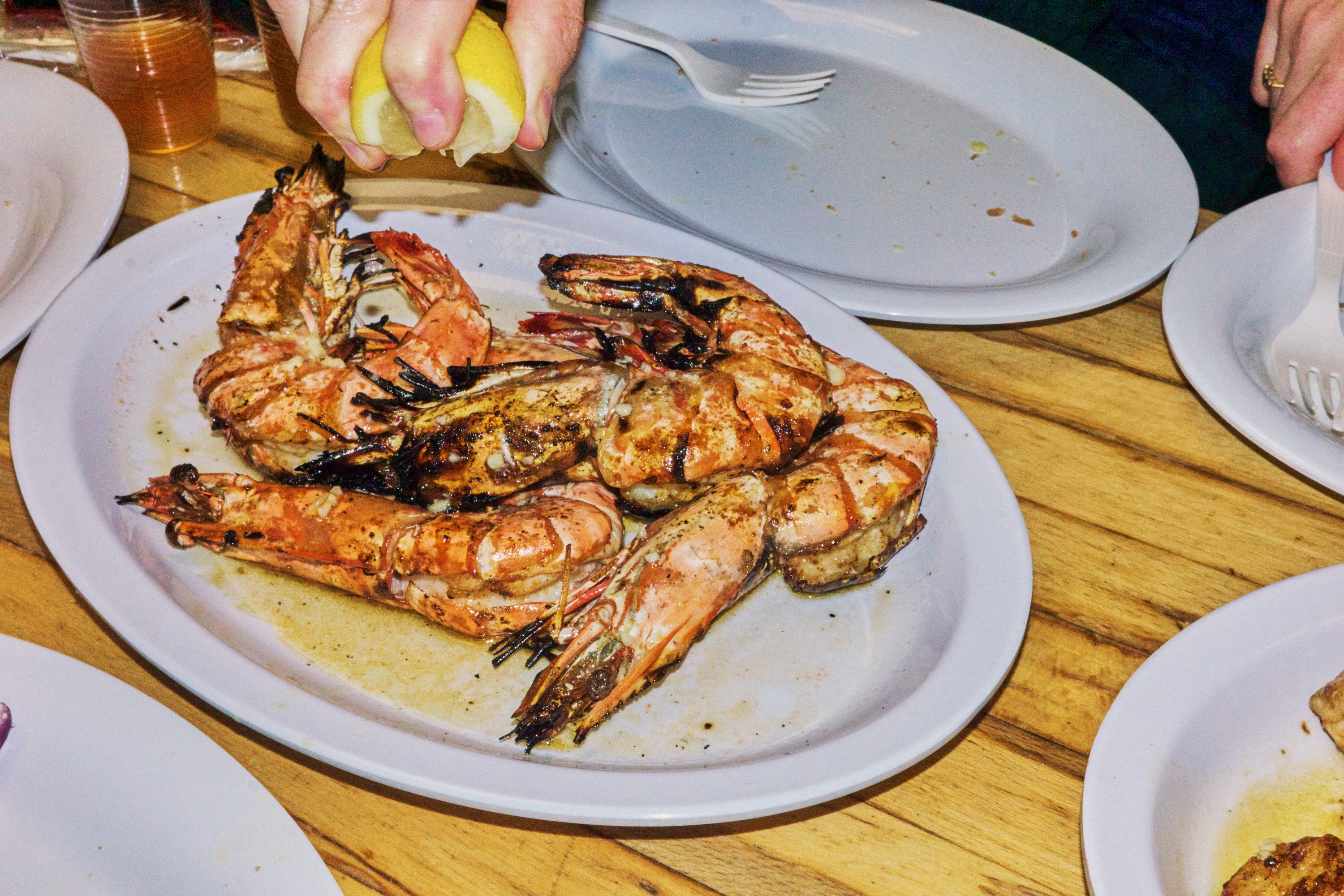 A hand squeezes a lemon wedge over a plate of grilled head-on shrimp.