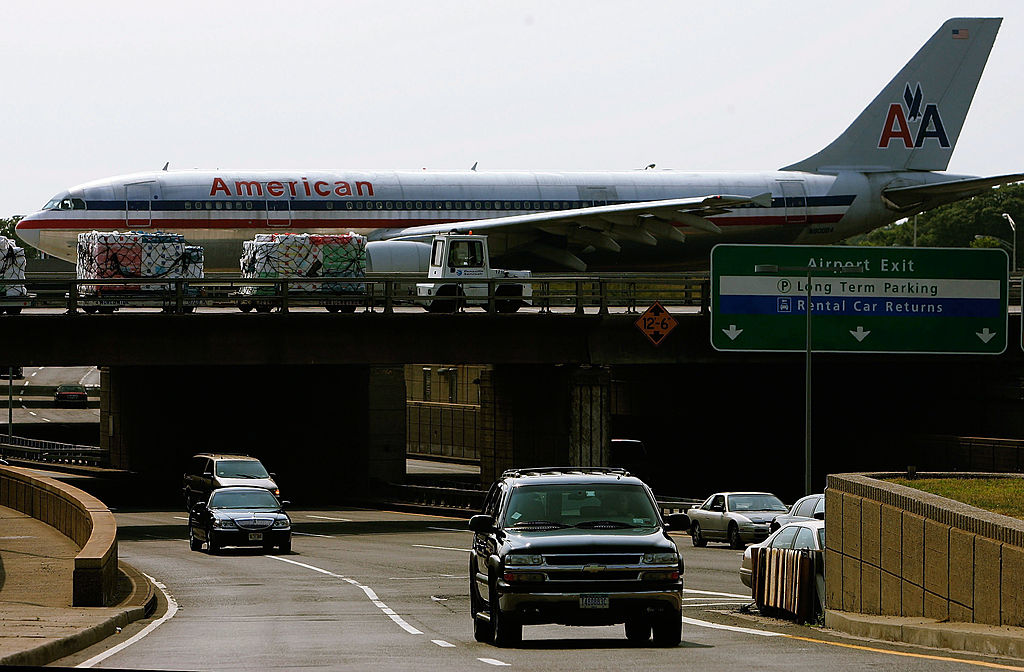 a passenger jet passes traffic overhead near JFK airport as trucks and other cars are on the road.