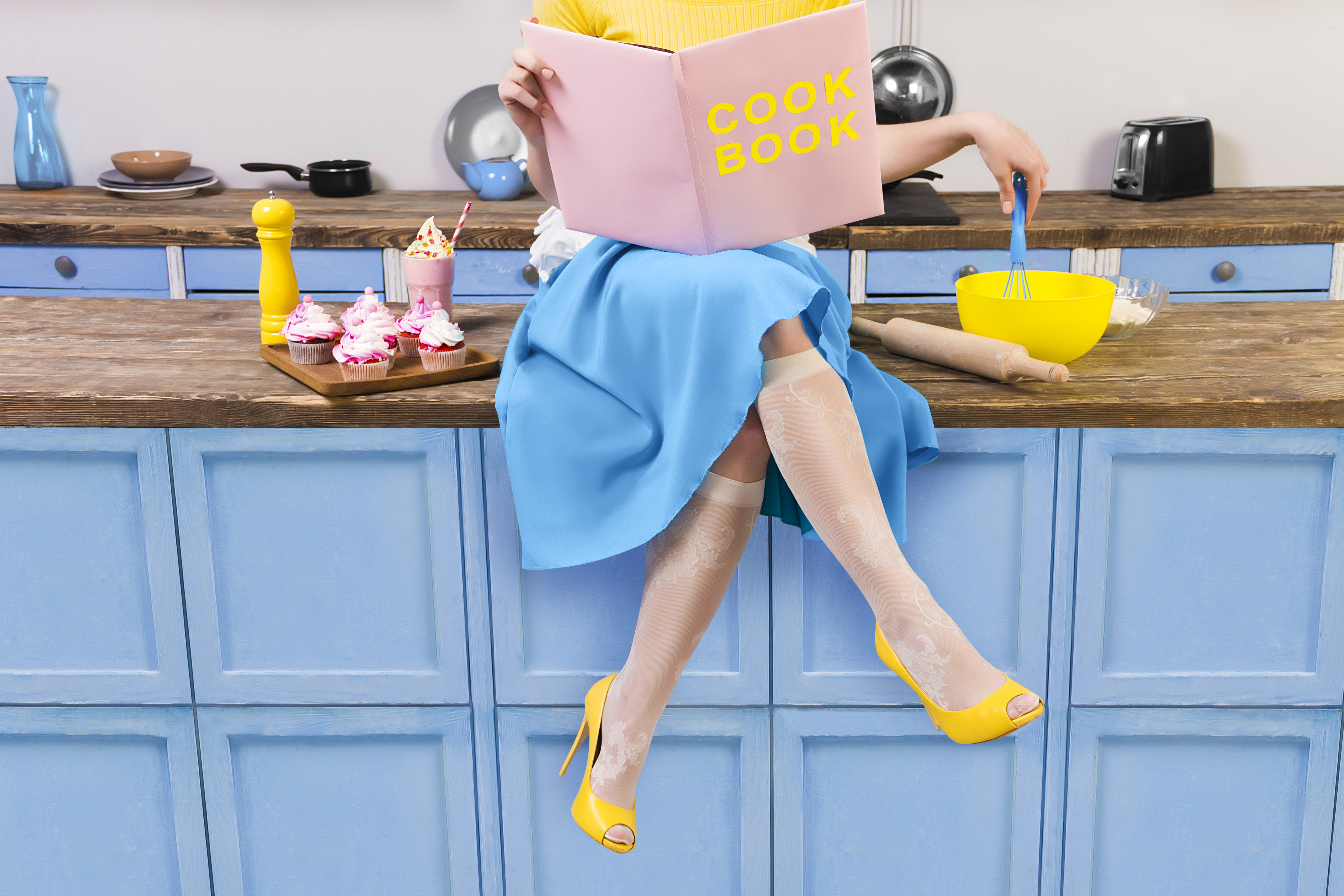 A woman sits on a kitchen counter with baking supplies holding a cook book.