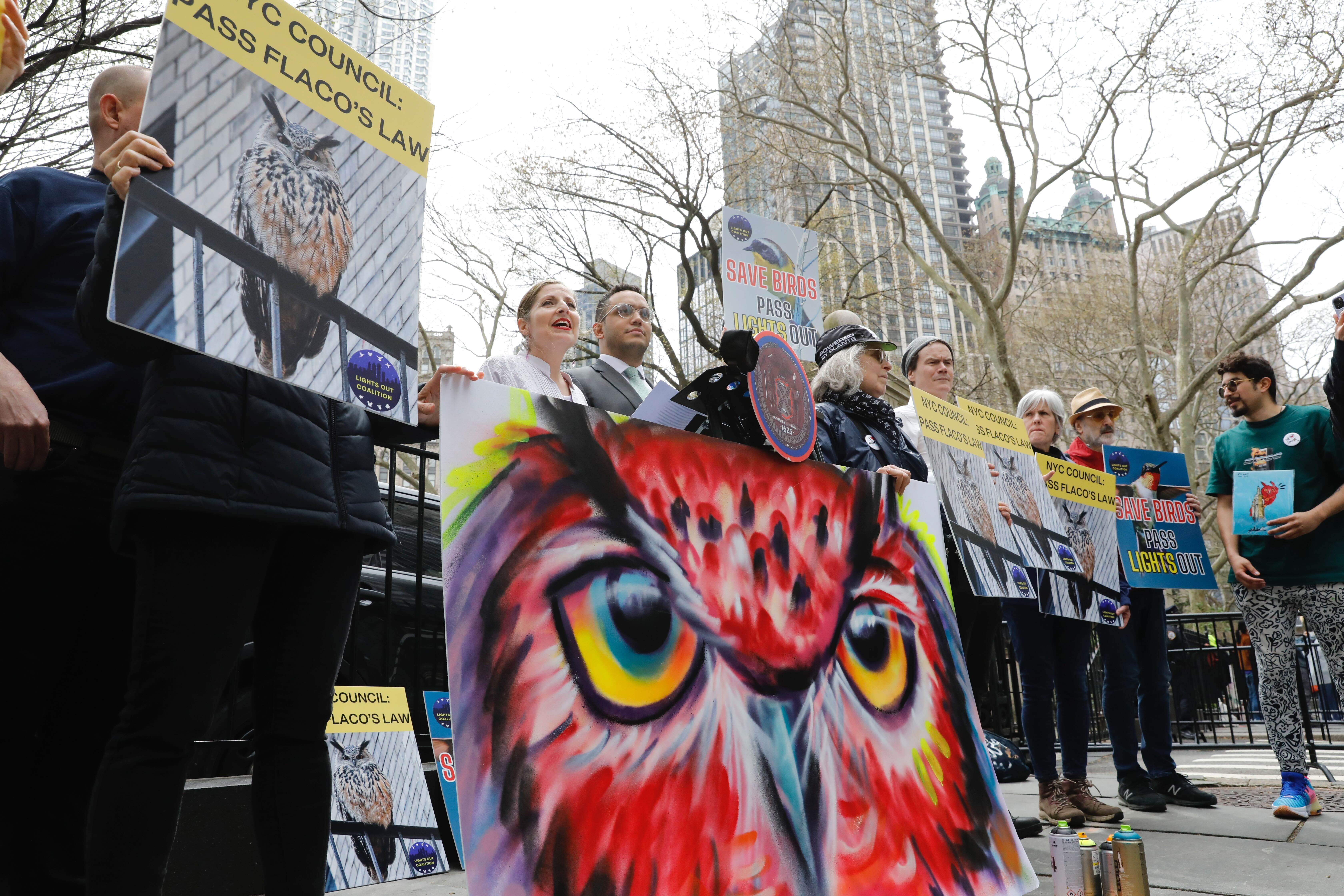 Politicians speak at an outdoor press conference alongside activists holding paintings of an owl.