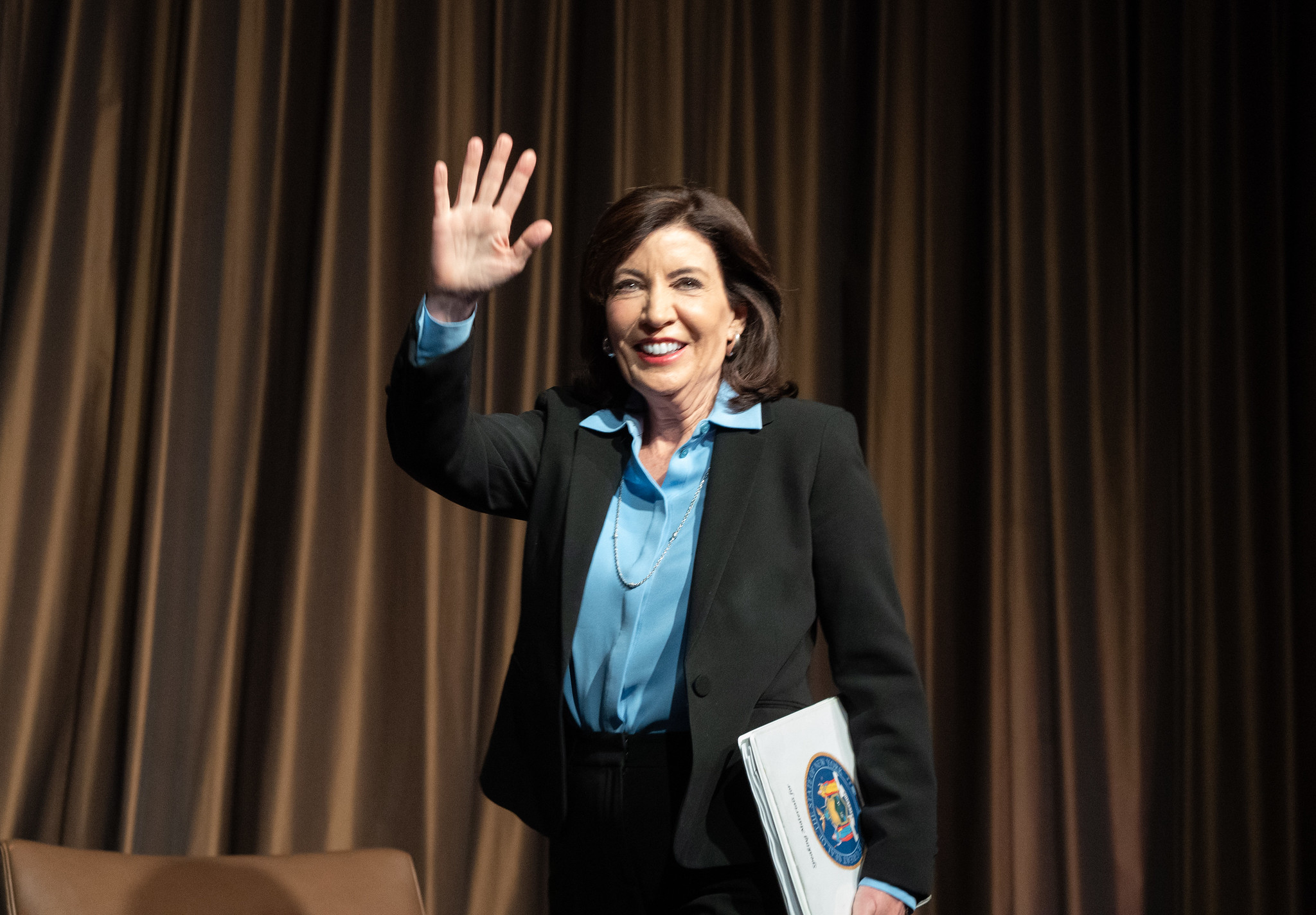 Gov. Kathy Hochul walks across a stage with a binder in one hand and waving with the other.