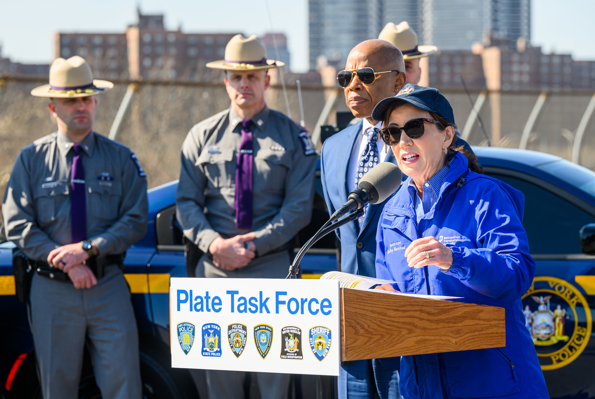 Gov. Kathy Hochul, wearing sunglasses and a blue windbreaker, stands alongside Mayor Eric Adams and State Troopers at a news conference on toll evaders.