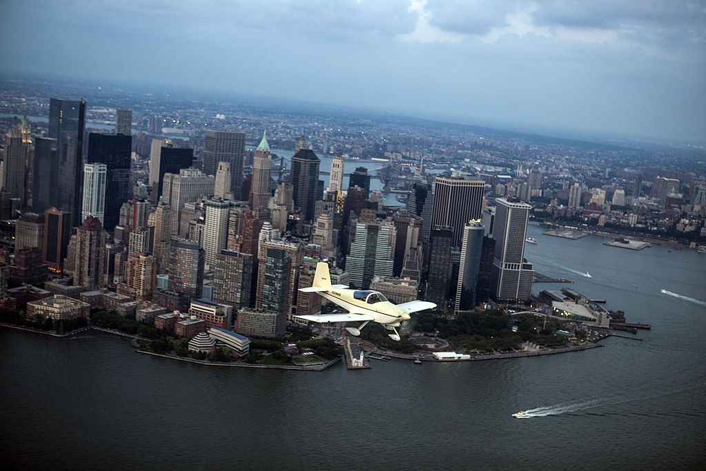 A cream colored prop plane flys over the Hudson River with Lower Manhattan in the background.