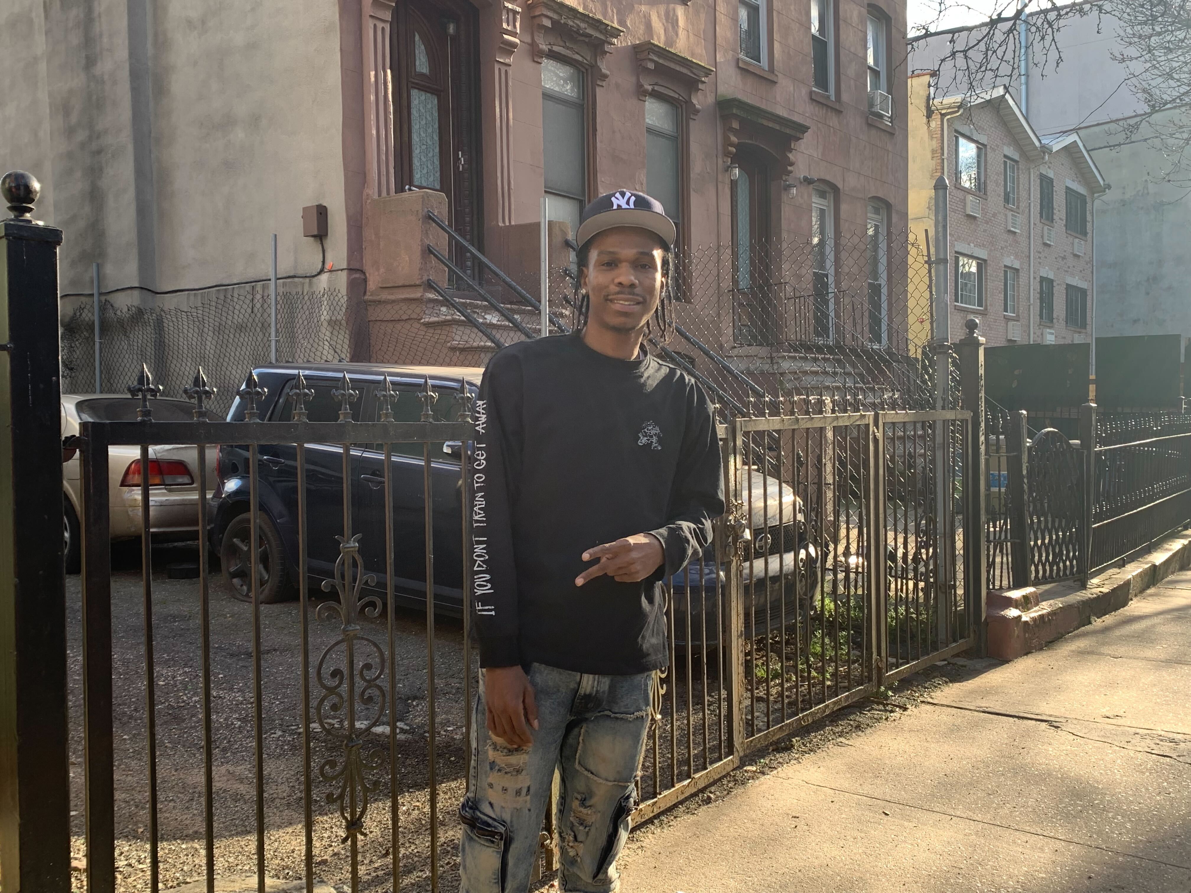 A man stands on the sidewalk with a brownstone in the background.