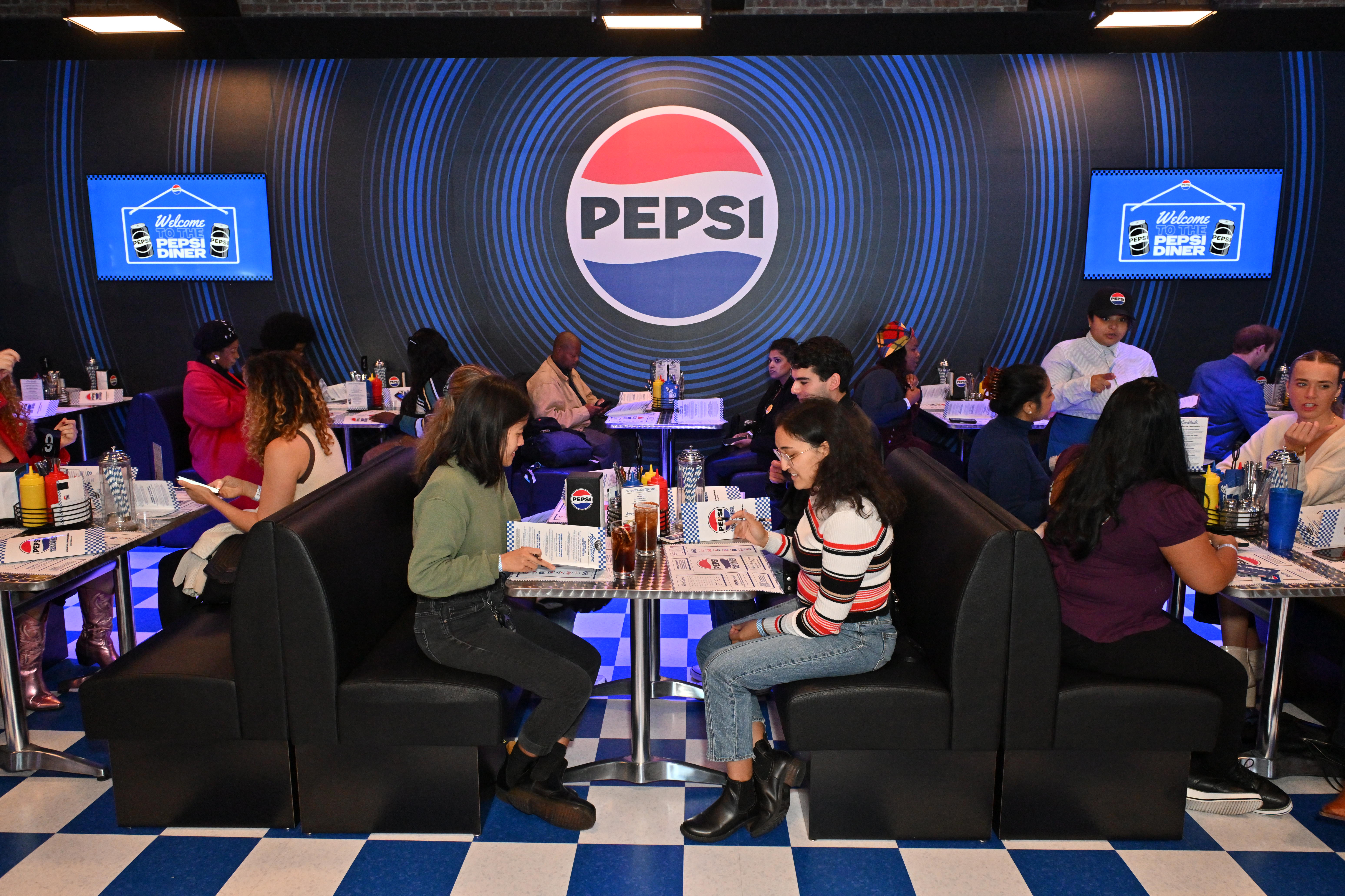 Two people sit at a diner with the word "Pepsi" behind them.