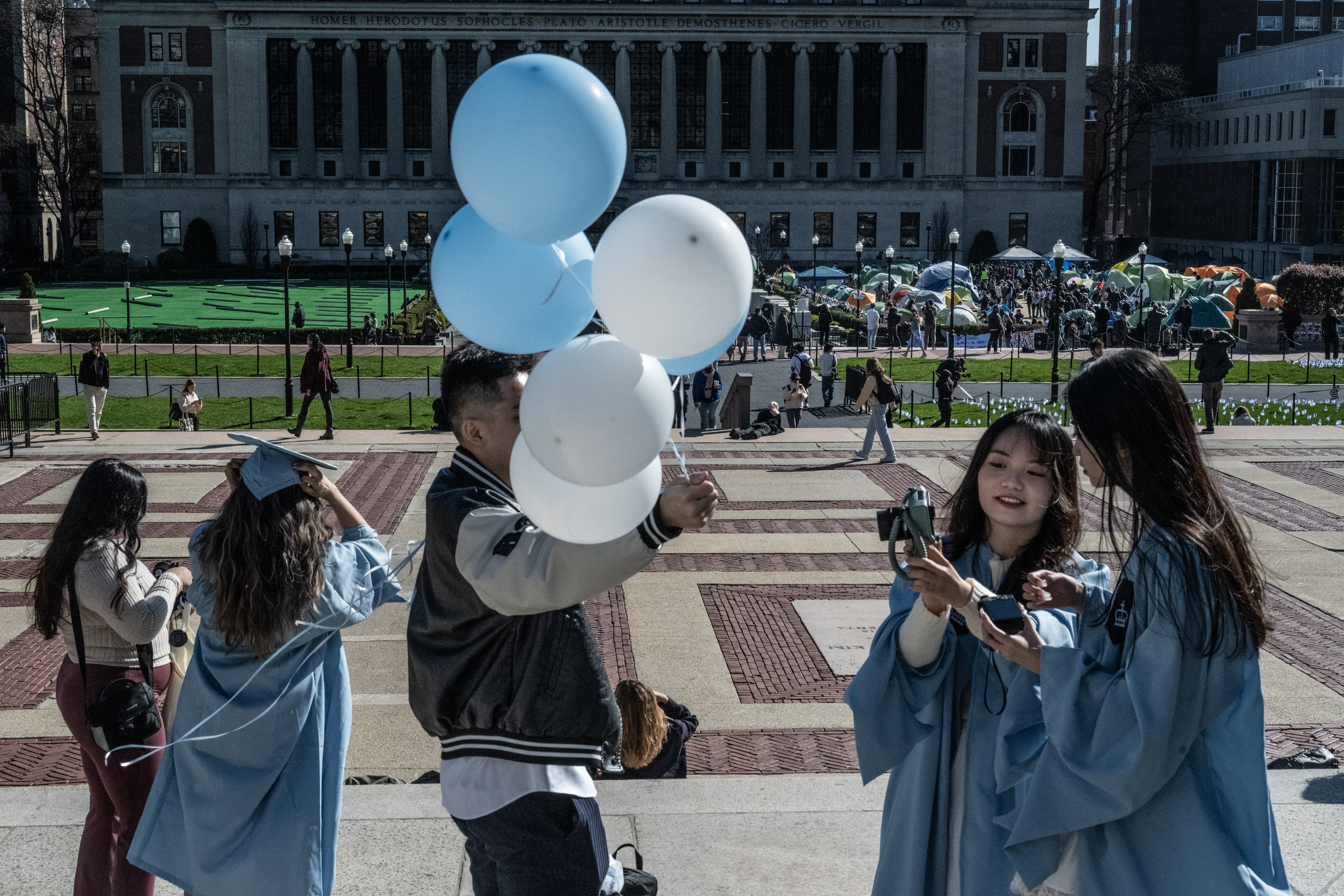 Students wearing graduation gowns at Columbia University in front of an encampment of protesters.