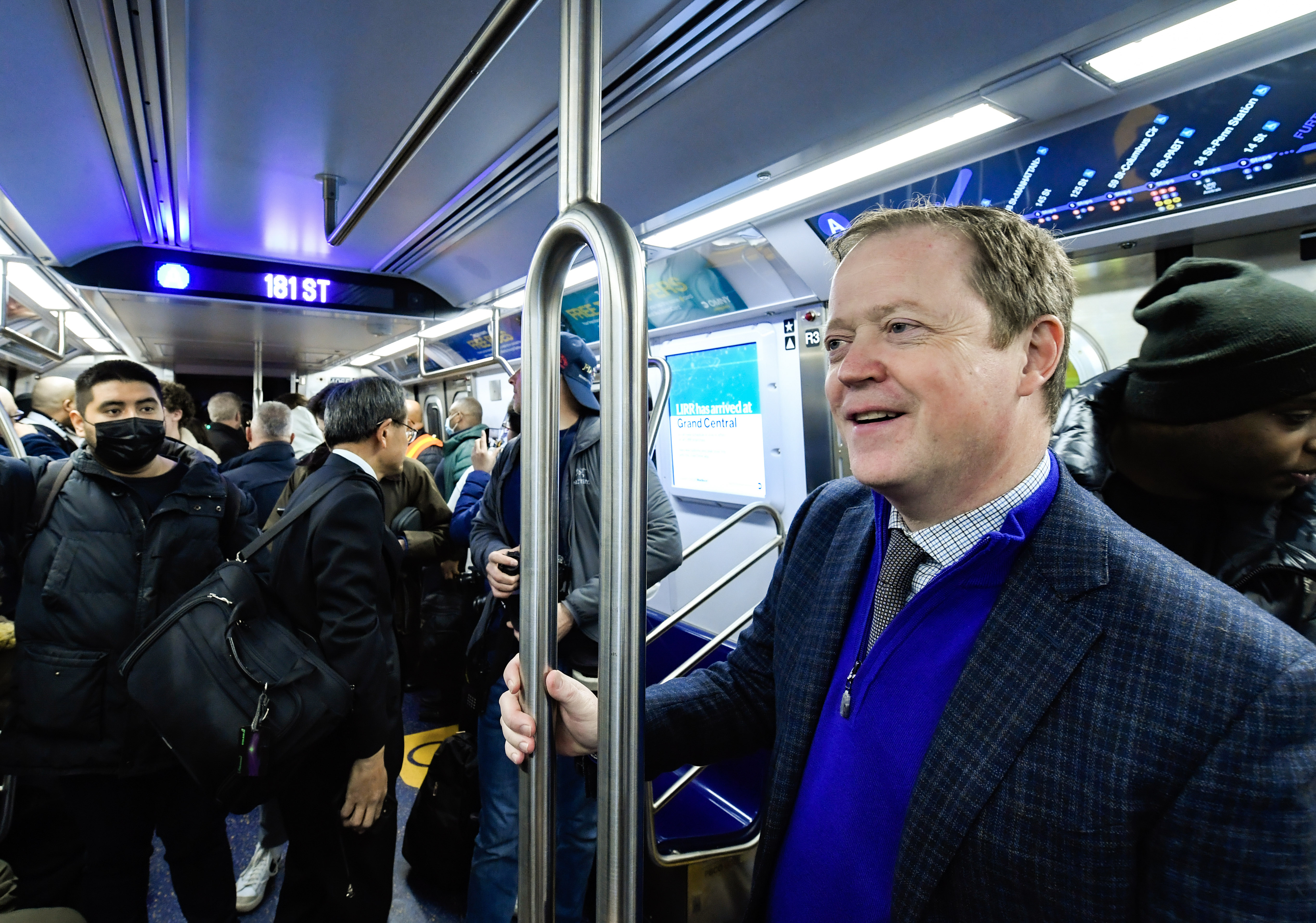 A man wearing a tie, quarter-zip jacket and blazer smiles on a subway train.