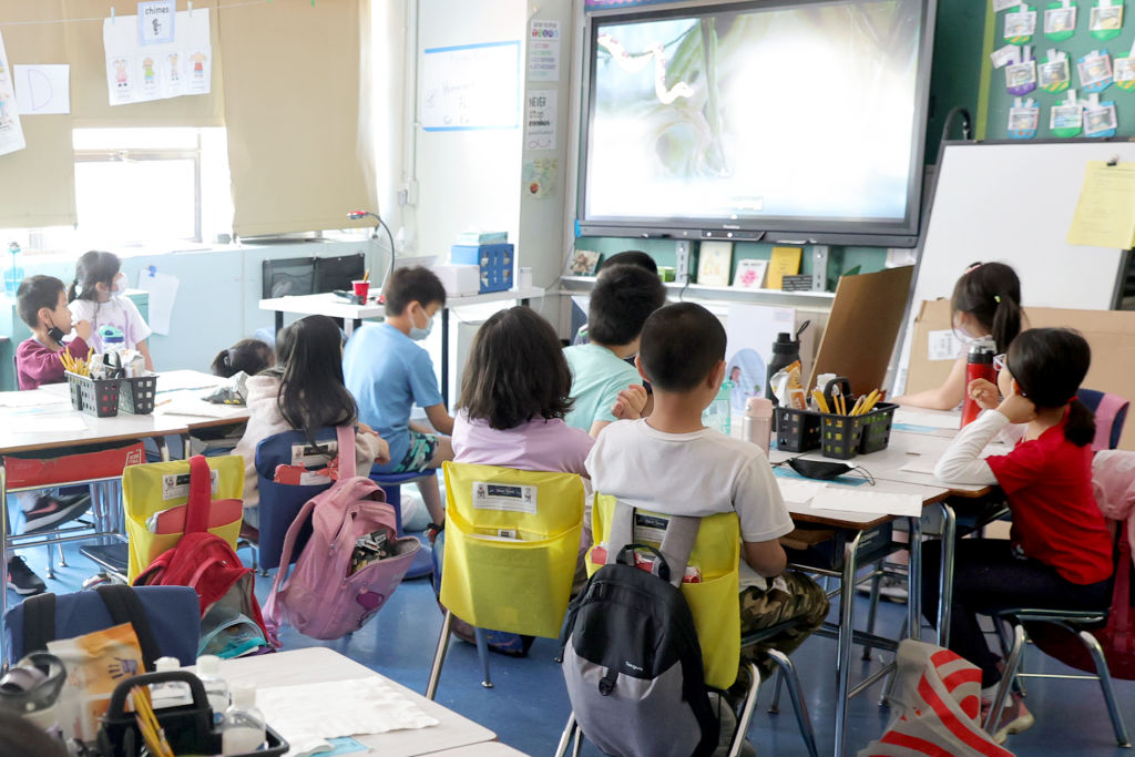 Young children sit at their desks in a classroom looking at the whiteboard.