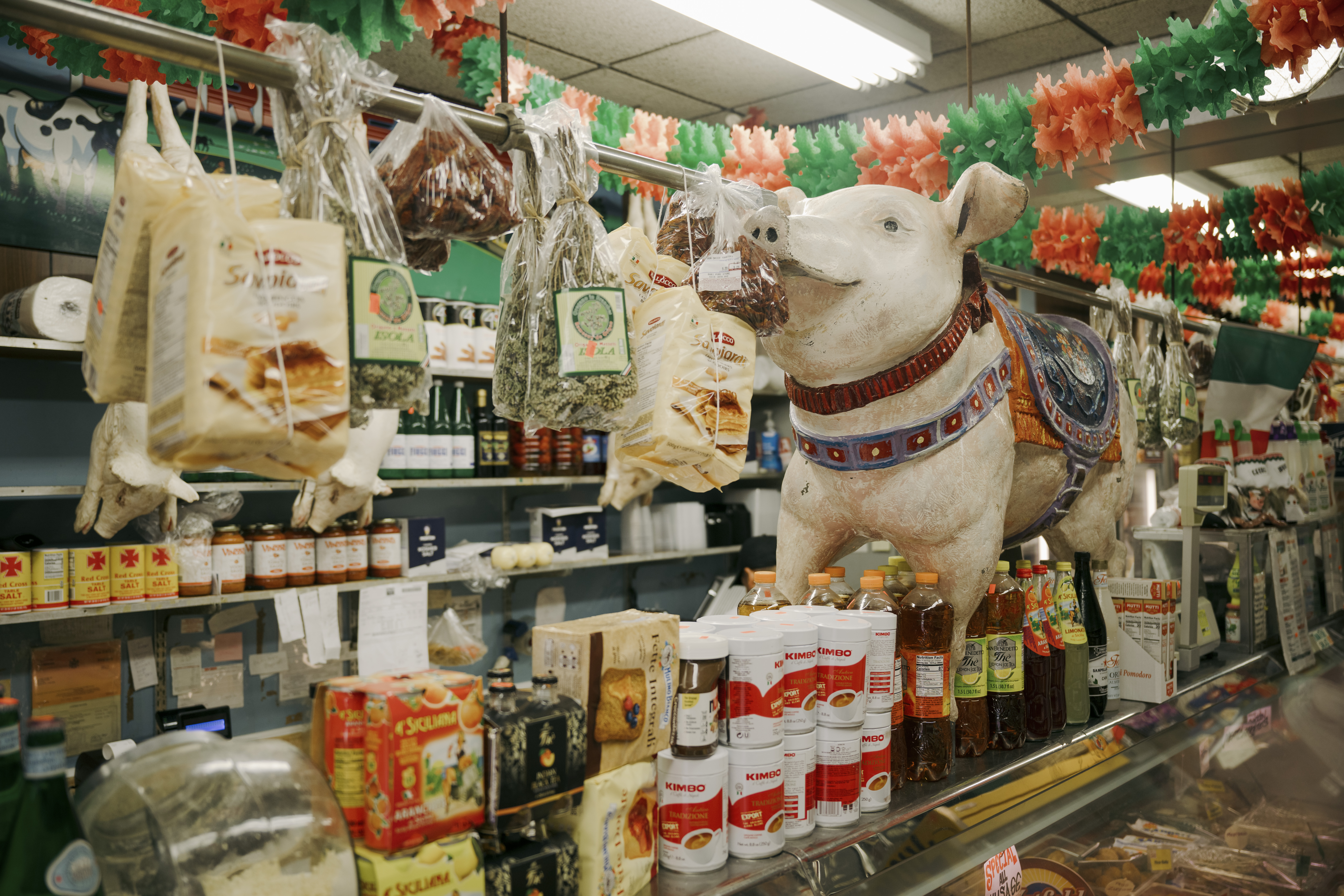 A deli counter is lined with canned goods and a figurine of a pig. Food items are hanging from a pole, and there are green and red decorations.