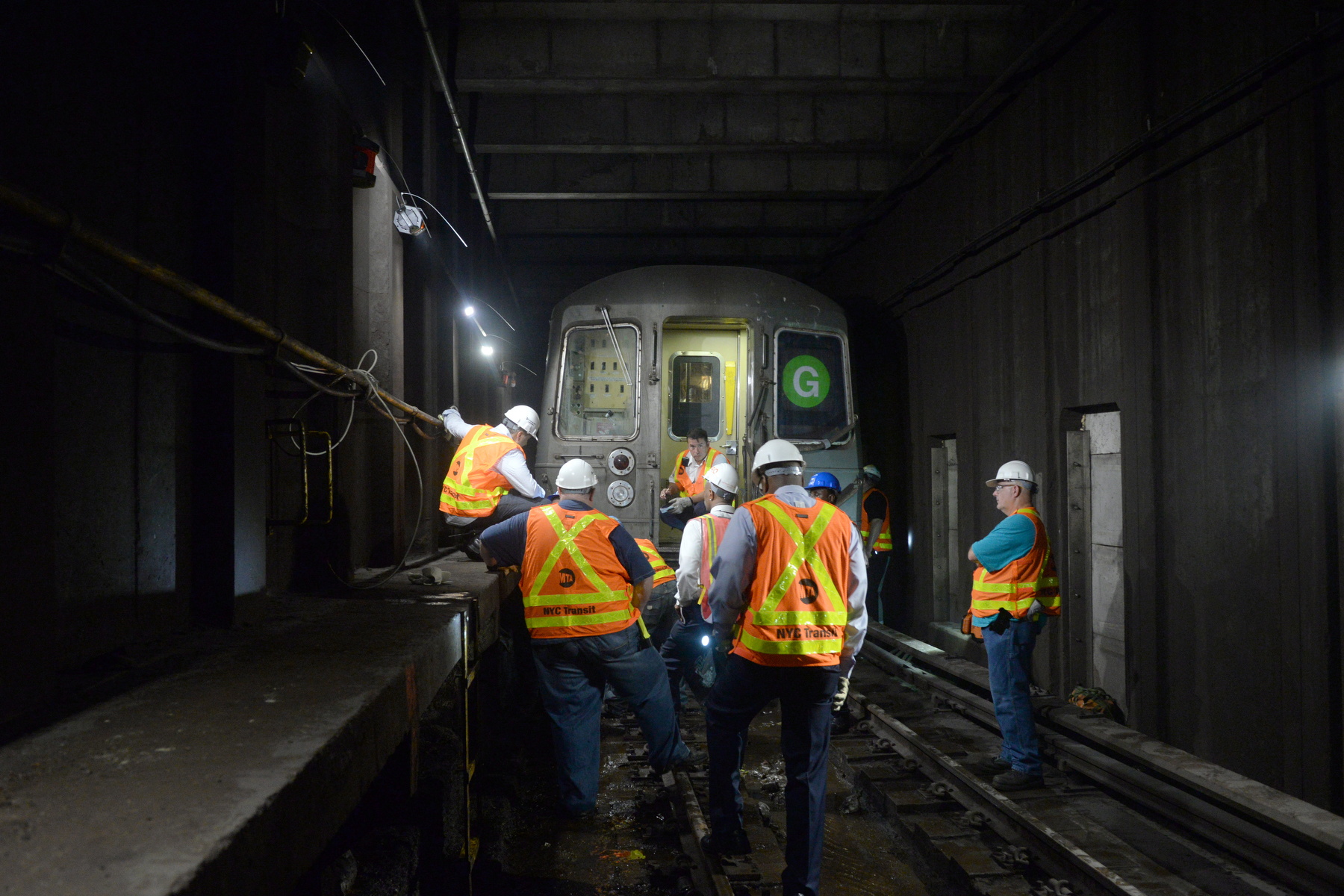 MTA workers in orange vests on the tracks in front of a G train.