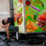 A delivery person sits with his head down and his hands clasped in sweltering heat. He is wearing green shorts and a black shirt.