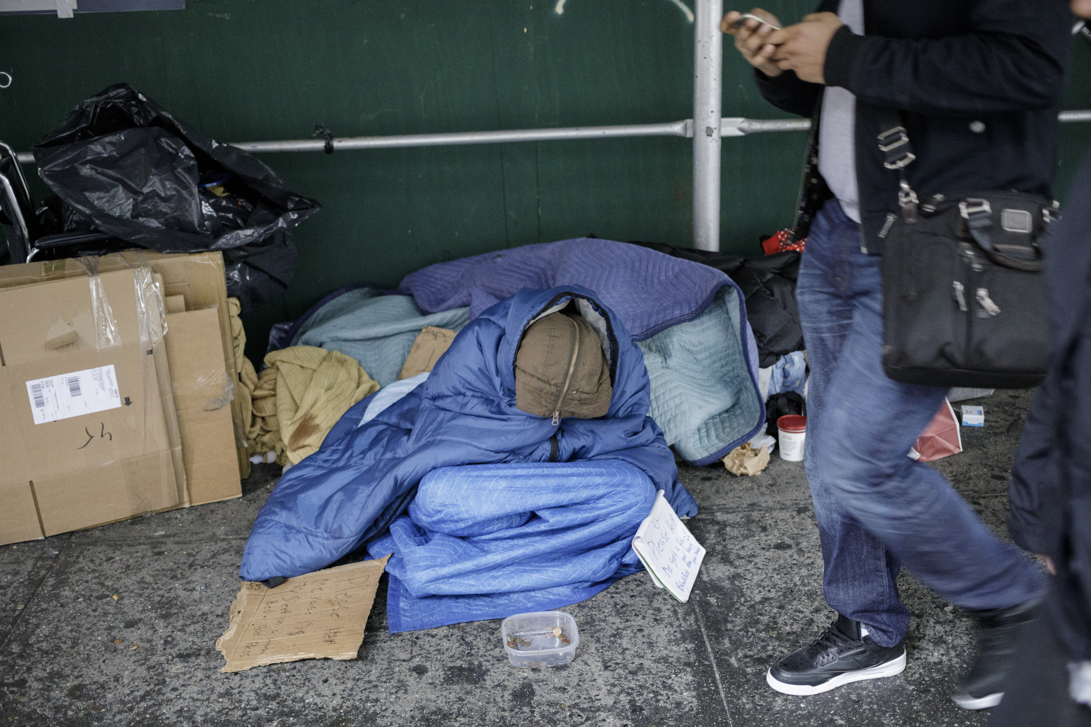 A homeless person sits wrapped with a blue moving blanket, while someone on their phone walks by.