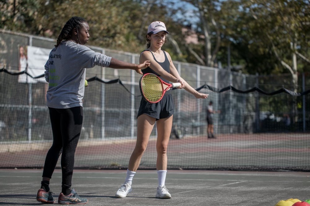 A woman gives pointers to another woman holding a tennis racket.